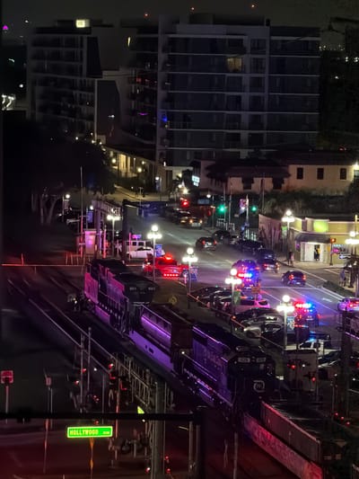 At night in Hollywood, Florida, a train passes by a scene with multiple law enforcement vehicles, including police cars with flashing lights. The train, a dark locomotive with purple lighting along its side, is on tracks in the foreground. In the background, a street is visible with numerous cars, some parked and others in traffic. Buildings line the street, including apartment complexes with illuminated windows and a lower-rise building with shops. The overall scene suggests an active law enforcement presence on a city street at night. A street sign reads "HOLLYWOOD BLVD."