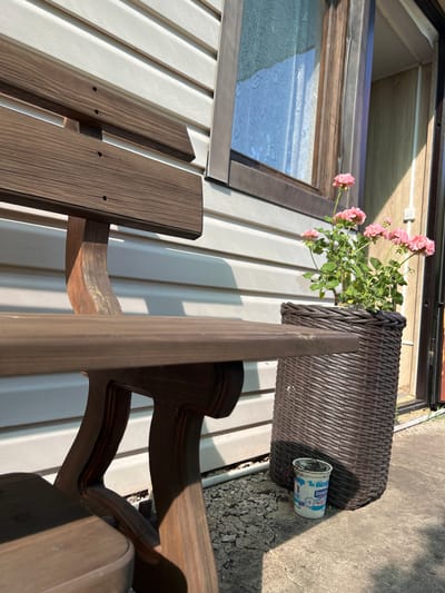 In the city of Tashelka, Russia, a wooden bench sits beside a building with white horizontal siding. In front of the building, a large, dark brown woven planter holds a flowering plant with pink blossoms. To the right of the planter, a small, white plastic cup with blue lettering is on the ground. The scene is illuminated by sunlight, casting shadows.