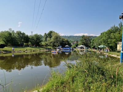 A sunny, clear day with a bright blue sky over a lake in German, Bulgaria. The lake is calm and reflects the surrounding trees and sky. Several people are visible by the water's edge, some fishing, others relaxing. Small boats are on the water. A large green umbrella and a white tent provide shade on the bank, and a sign with Bulgarian text is posted on a blue pole. Tall green trees and a hill covered in greenery form the background. A few power lines stretch across the upper part of the frame.