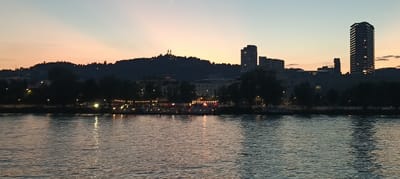 The image shows a panoramic view of the Danube River in Linz, Austria, during sunset. The sky displays a gradient of colors, from soft orange and pink near the horizon to pale blue at the top. On the far bank of the river, a silhouetted hillside is visible, topped with two prominent church spires, likely the Pöstlingberg Basilica. Below the hill, the city skyline is illuminated with lights. Several boats are docked along the riverbank, adorned with string lights, suggesting a lively evening atmosphere. In the foreground, the river's surface reflects the twilight sky and the city lights, creating shimmering patterns. The overall scene captures the serene beauty of Linz at dusk.