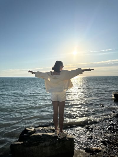 A person stands on a rock facing away from the camera, with arms outstretched. The person is wearing a light-colored jacket and white shorts, with white sneakers. The sun is setting over a body of water, casting a bright reflection on the waves. The background shows a clear blue sky with a few wispy clouds. The ground around the person is rocky and leads down to the water. The location is the coast of Lake Sevan in Supsekh, Russia.