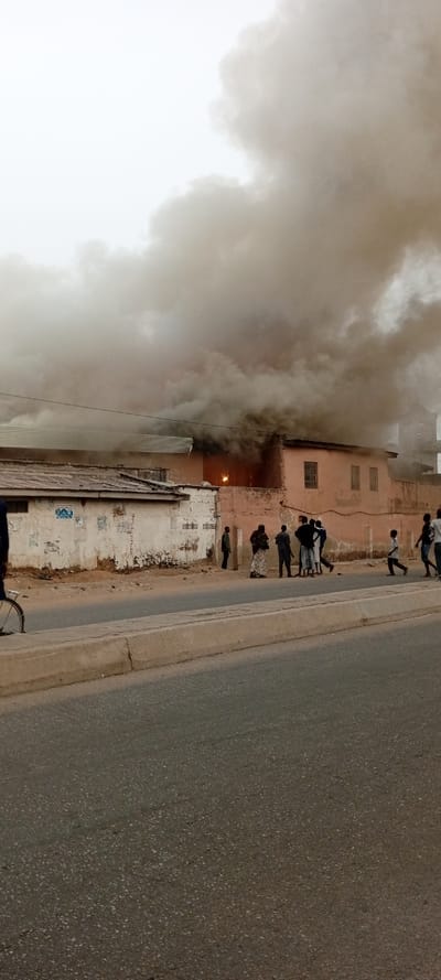 The image captures an outdoor scene dominated by a building fire, with thick, dark smoke billowing into the sky. The setting appears to be a street or roadside in an urban or semi-urban environment.
In the mid-ground, a light brown or peach-colored building is ablaze. Large, dense plumes of dark gray and black smoke rise from the structure, almost entirely obscuring the upper portion of the building and a significant part of the sky. An intense orange glow of flames is distinctly visible within an opening or window of this burning building, confirming the active fire. To the left of the burning structure, another building with a corrugated metal roof is partially visible. Its wall, painted in a lighter, possibly off-white or cream color, shows signs of wear and tear, including peeling paint and faint remnants of posters or advertisements, though no specific text is legible.
Several people, including adults and children, are gathered on the dirt shoulder or sidewalk in front of the buildings, observing the fire. They are dressed in casual clothing. Some individuals are standing still, watching the event unfold, while others, particularly on the right side of the group, appear to be moving, possibly walking or running. On the far left of the image, the front wheel and part of the frame of a bicycle are partially visible.
The foreground consists of a paved asphalt road, separated from the dirt shoulder by a low, light gray concrete curb. The lighting suggests it is daytime, but the heavy smoke and possibly overcast conditions create a somewhat dim and diffused atmosphere. The scene depicts a dramatic event, with observers drawn to witness the unfolding fire.