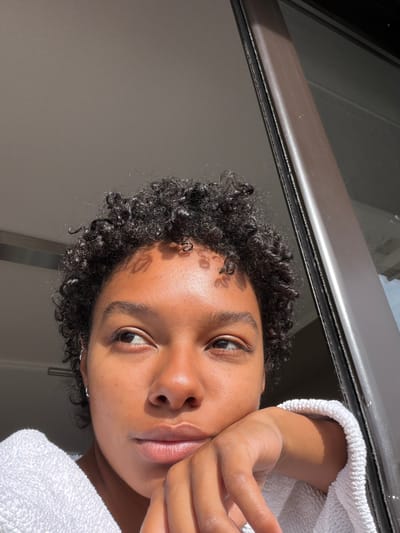 A person with dark curly hair and brown skin is close to the camera, positioned near a window. The person is wearing a white towel. The window is visible behind the person, with a metal frame. Light casts shadows on the person's face. There is no information in the image to determine if the location is in Champs-sur-Marne, France.