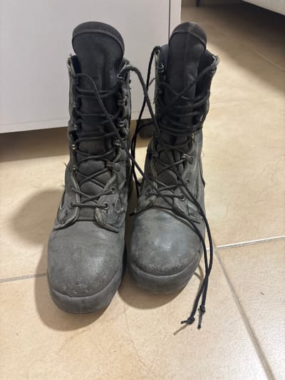 The image displays a pair of well-worn, dark grey military-style combat boots standing upright on a light brown tiled floor in Maale Adumim, Palestinian Territory. The boots feature black laces loosely threaded through multiple metal eyelets, with the ends untied and resting on the floor. The material, appearing as a combination of leather or synthetic equivalent on the lower sections and textile on the upper shafts, shows visible scuffs, dirt, and general wear, particularly on the toe caps. A white, opaque surface, likely a cabinet or wall, is partially visible in the background. No individuals or active interactions are present in the scene.