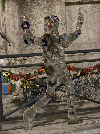 The image shows a life-size human-shaped sculpture constructed from numerous CDs, which reflect light and create a shimmering effect. The sculpture stands upright, with its arms raised. It is positioned outdoors, in front of a stone wall and behind a metal railing. A garland of artificial red and yellow flowers is draped across the railing. The ground appears to be paved. The overall lighting suggests it may be evening or nighttime, with artificial illumination highlighting the CDs. There are no people present in the immediate vicinity of the sculpture. The background includes rough-textured stone walls and some greenery. There are no visible signs or indications that this sculpture is located in Vienne, France.