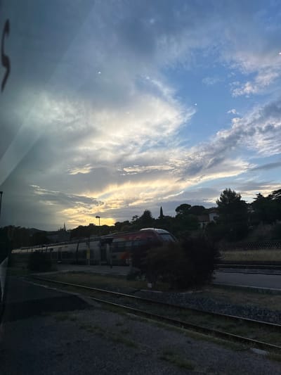 The image shows a train stopped at a station with a dramatic sky overhead. The sky is filled with clouds, some of which are illuminated by the setting sun, creating a warm glow. The train appears to be modern, with a silver and red exterior. It is stopped on a track, with the platform visible to the right. In the background, there are trees and buildings, suggesting an urban or semi-urban setting. The overall impression is one of calm and peacefulness, with a hint of the dramatic power of nature. The location is the train station in Alès, France, a town in the Occitanie region of southern France.
