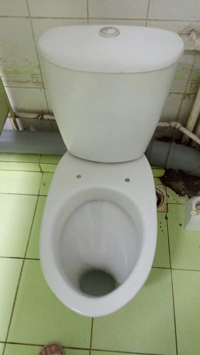 A top-down, slightly angled view shows a white ceramic toilet in a small, tiled room. The toilet tank sits atop the bowl, and the flush mechanism is visible on the top of the tank. The toilet bowl itself appears to have two small holes on its rim, resembling eyes, and the interior of the bowl contains a dark liquid. The floor is covered in light green square tiles, with visible grout lines. To the right of the toilet, pipes and a dark, grimy area are present near the wall. A white object, possibly a toilet paper holder, is mounted on the wall to the right of the toilet. In the lower left corner, the foot of a person wearing a sandal is visible. The overall impression is that of a utilitarian, possibly older, bathroom. The image is not tagged with a specific location in Tomsk, Russia.