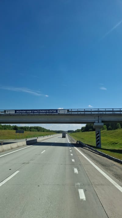 The image shows a view from the driver's perspective on a highway in the city of Sokur, Russia. The highway is paved with asphalt and has multiple lanes separated by white lines. There are concrete barriers on the sides of the road. A pedestrian overpass spans the highway, with a banner on it that reads "ЖЕЛАЕМ ВАМ СЧАСТЛИВОГО ПУТИ!" which translates to "We wish you a happy journey!". The sky is clear blue with a few wispy clouds. The surrounding environment consists of green fields and trees. A truck is visible on the highway in the distance. The overall scene depicts a typical highway landscape on a sunny day in Russia.