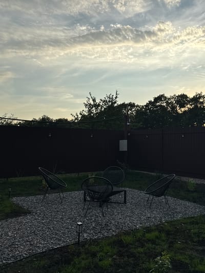 This is a vertical, outdoor shot of a backyard area at dusk. The scene features a dark, slatted fence running horizontally across the mid-ground. To the left of the fence, the sky is filled with wispy, elongated clouds illuminated by the setting sun, creating a dramatic, streaky effect. Trees with dark foliage line the top of the fence, silhouetted against the sky.
In the foreground, a pathway of dark grey gravel leads diagonally from the bottom right to the top left of the frame. On this path, there are two black, wire-frame chairs with rounded backs and a small, low black table between them. Two small, rectangular garden lights are visible along the edge of the gravel path. To the right of the gravel path, a strip of dark green grass is visible, with some lighter green foliage growing in it. Another garden light is planted in the grass near the bottom edge of the frame.
The overall mood is tranquil and slightly melancholic, fitting for the twilight hour. The light filtering through the clouds suggests a cool evening. The setting appears to be a private residence. There is no visible text in the image.