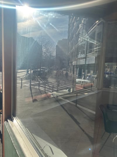 The image depicts a sunlit urban street scene in Grenoble, France, viewed through a large window, which exhibits strong glare and reflections. The foreground shows the lower section of a window frame and reflections of an interior space, including a dark chair on a tiled floor.

Outside, a paved sidewalk runs parallel to the viewer, featuring a bike rack with several bicycles parked. Beyond the bike rack, a crosswalk with white stripes extends across the street. Dark, vertical bollards are positioned along the sidewalk near the crosswalk. Further down the sidewalk, an orange planter with soil is visible.

The street itself is lined with parked cars on the left. Multiple pedestrians are visible on the distant sidewalk; at least two figures are walking away from the viewer on the right side of the street, and another less distinct figure is further down the street on the left.

Multi-story buildings, consistent with urban European architecture, line both sides of the street, featuring numerous windows and some balconies. Bare trees, indicative of winter or early spring, are interspersed along the street. The sky is clear blue with some faint wispy clouds and contrails. The sun, positioned in the upper left, casts significant glare and lens flare across the window, creating strong shadows from street objects onto the pavement and across the interior floor visible in reflection.