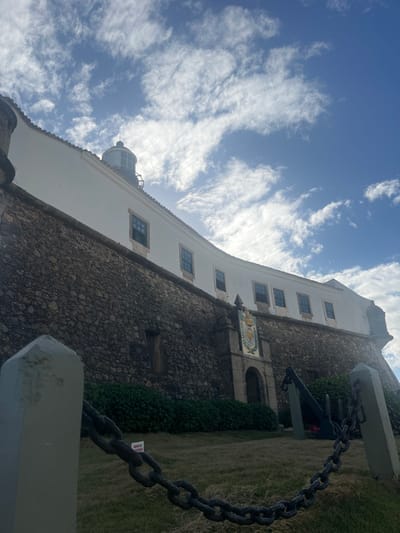 The image displays a low-angle view of the Santo Antônio da Barra Fort, also known as Farol da Barra, situated in Salvador, Bahia, Brazil.
In the foreground, a dark metal chain barrier extends across the bottom, connected between two light-colored, pyramidal-topped posts. The ground slopes upwards, covered in dry, light green grass, with a line of dense green bushes bordering the fort's base. To the right of the fort's central entrance, a large black anchor with a chain rests on the grass.
The fort's lower structure is a robust, dark-colored stone wall constructed from irregular masonry. Centrally positioned on this wall is a decorative arched stone entrance, topped by an ornate coat of arms or crest. Above the stone wall, a white-painted building with a gently curving facade features multiple evenly spaced rectangular windows with dark frames. At the highest point of this white structure, towards the left, the cylindrical lantern room of the lighthouse is visible, capped by a dark, conical roof and finial.
The background consists of a bright blue sky filled with scattered, textured white cumulus clouds. No individuals are present in the scene.