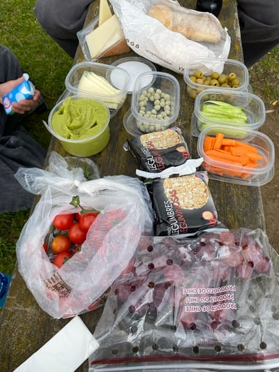 The scene shows a picnic spread on a weathered wooden surface, possibly a picnic table. The food includes a baguette, olives, sliced cucumbers and carrots, green coated spheres, rice cakes, grapes in a clear plastic bag labeled "PRODUCT OF CHILE", tomatoes in a plastic bag, sliced cheese, and green dip. A person is visible in the lower portion of the image, wearing grey pants and holding a small blue and white carton. A napkin is also visible. The food items are a mix of packaged and fresh items, suggesting a casual outdoor meal. Without other visual cues, the location in Villaviciosa, Spain is not identifiable.
