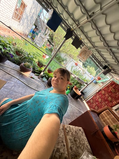 A woman in a blue floral dress is pictured in an outdoor, covered patio area. The setting appears to be a backyard with a tiled floor and a corrugated metal roof overhead supported by metal beams. Laundry is hanging on a line, including dark t-shirts and a patterned cloth with a tiger design. Pots of plants and flowers are visible throughout the scene, some hanging and others on the ground. A garden hose lies coiled on the floor.
In the background, a brick building with a window can be seen, along with a lush green garden area. Further back, there's a glimpse of a greenhouse-like structure and some trees. To the right of the woman, a dark wooden piece of furniture, possibly a dresser or table, is present, topped with a red patterned fabric and a potted plant. Another piece of furniture, upholstered in a floral fabric, is also visible.
The lighting suggests it is daytime, likely sunny given the bright illumination and shadows. The overall atmosphere is relaxed and domestic, possibly indicating a summer day. The woman's expression is neutral as she looks directly at the camera. No visible text is present in the image. The location context is Nizhne-Chufichevo, Russia.