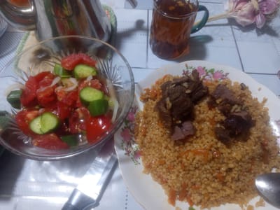 This is a medium-angle, eye-level shot of a meal set on a patterned tablecloth. The scene appears to be indoors, likely in a home, given the casual presentation and the presence of common household items.
The main subjects are food and drink. On the left, a clear glass bowl contains a fresh salad made of chopped tomatoes, cucumbers, and sliced white onions. To the right of the salad, a plate is filled with pilaf, a rice dish mixed with carrots and topped with pieces of dark, cooked meat. A glass mug filled with a dark amber liquid, presumably tea, sits behind the salad bowl. In the background, a bouquet of garlic is tied together with a string. A metal teapot is partially visible in the upper left corner, and a metal spoon rests on the edge of the pilaf plate. The setting is a table covered with a white and blue patterned tablecloth.
The image captures a moment of dining, suggesting a meal is either about to be eaten or has just been served. There are no people visible in the image. The lighting suggests it might be daytime, but the exact time of day is not discernible. The overall mood is one of a simple, home-cooked meal. No visible text is present in the image. The location context provided is Razzakov, Kyrgyzstan, which aligns with the typical cuisine depicted.
