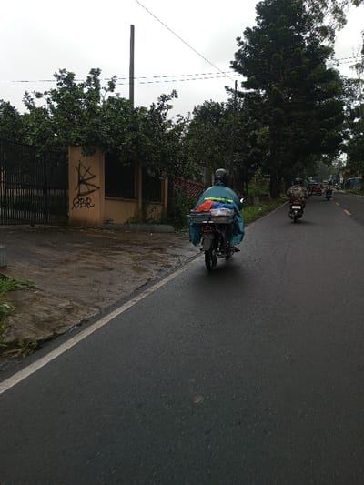 The image displays a street scene in the city of YonPhoogla, Bhutan, under an overcast sky, with damp conditions suggesting recent precipitation. In the immediate foreground, a wet asphalt road features a solid white line along its left edge. A person, wearing a dark helmet and a blue-green rain poncho, operates a motorcycle, moving away from the viewer in the left lane. The motorcycle's rear is equipped with a dark grey carrier holding various items, including red, blue, white, and green plastic bags, and other packaged goods. Further down the road, a second individual on a motorcycle, wearing patterned clothing and a dark helmet, also travels away from the camera, followed by additional motorcycles in the distance. To the left of the road, a wet concrete surface extends towards a light tan architectural structure, possibly a wall or gate pillar, which bears dark graffiti including the letters "OBR" and abstract marks. Behind this structure, a dark metal fence or gate is partially visible, obscured by dense green foliage. A tall, slender utility pole stands behind the gate. The right side of the road is bordered by a line of tall, dark green trees, and the road itself curves gently to the right as it recedes. Overhead, multiple utility lines are visible against the pale sky.
