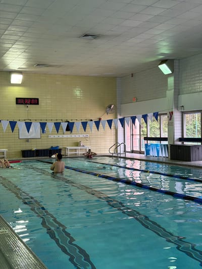 This image depicts an indoor swimming pool in Bethesda, United States of America. The pool is divided into lanes by lane markers. Several individuals are in the pool, with one person standing and others swimming. The pool's perimeter includes walls tiled in white and yellow. A digital timer is visible on the wall. There are windows and pool equipment visible near the pool. Decorations such as hanging pennants are present along the walls.