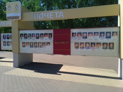 This media file is a medium-shot, eye-level photograph of an outdoor display board in Otradny, Russia. The display board, which is part of a larger structure, features multiple rows of framed portraits of individuals. Above the portraits, large white letters spell out "ПОЧЕТА" on a yellow banner, meaning "HONOR" in Russian. To the left of this banner, a coat of arms is visible. Below the "ПОЧЕТА" banner, there is a red section with white text that reads "ПРЕДСТАВИТЕЛИ ПРЕДПРИЯТИЙ," which translates to "REPRESENTATIVES OF ENTERPRISES."
The portraits are arranged in rows and columns, with each person having a name printed below their image. The individuals appear to be men and women of various ages, dressed in formal or business attire. The background of the display board is white. The scene is set outdoors on a paved area, likely a sidewalk or plaza, with a shadow cast from the display structure. In the background, lush green trees are visible against a bright blue sky, suggesting a sunny day. The overall impression is one of a public space recognizing esteemed individuals or businesses within the community. The time of day appears to be daytime, given the strong sunlight and shadows.