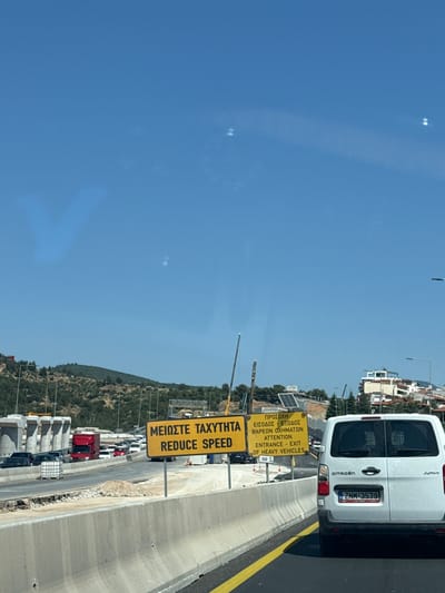 This media file captures a street scene in Pylaia Municipal Unit, Greece, likely during the daytime given the bright sunlight and clear blue sky. The perspective is from a vehicle, looking forward and slightly to the right, with a strong sense of motion implied by the blur of the road markings and the angle of the shot.

The primary subjects are a white Citroen Jumpy van and several road signs. The van, with the license plate ZNM-3579, is positioned on the right side of the road, partially obscuring the view of other vehicles behind it, which include cars and a red truck. The road itself appears to be a multi-lane highway or a similar type of thoroughfare, with concrete barriers and yellow lane markings visible.

Several yellow road signs are prominently displayed. One sign, written in both Greek and English, reads "ΜΕΙΩΣΤΕ ΤΑΧΥΤΗΤΑ REDUCE SPEED". Below this, another sign states "ΠΡΟΣΟΧΗ ΕΙΣΟΔΟΣ - ΕΞΟΔΟΣ ΒΑΡΕΩΝ ΟΧΗΜΑΤΩΝ ATTENTION ENTRANCE - EXIT OF HEAVY VEHICLES" along with a 50m indicator. The presence of these signs suggests an upcoming junction or a change in road conditions, specifically for heavy vehicles.

In the background, a hilly landscape covered in trees is visible, with buildings sparsely dotting the slopes, hinting at a semi-urban or suburban environment. The overall atmosphere is one of routine travel, with the focus on road safety and traffic management. The weather appears to be clear and sunny.