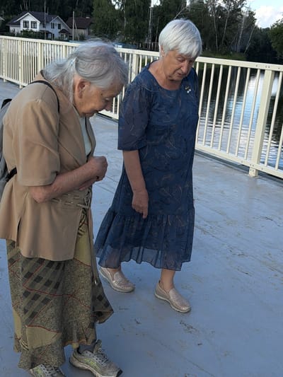 Two elderly women are standing on a bridge with a white railing. The woman on the left is wearing a tan jacket over a white shirt and a patterned skirt, and has gray hair. The woman on the right is wearing a blue patterned dress and has short white hair. They are both looking down. The background shows a body of water and trees, with houses visible in the distance. The bridge appears to be in a park or recreational area in the city of Udelnaya, Russia.