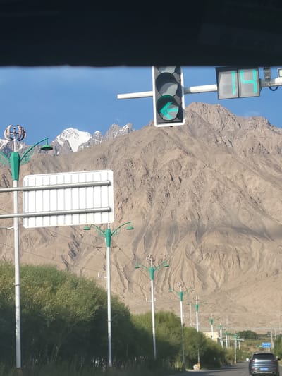 The image shows a view of a road in Tashkorgan, China, with a large mountain range in the background. A traffic light is visible in the foreground, displaying a green pedestrian symbol and a countdown timer showing "14" seconds. There are also several lampposts with decorative tops and a large white rectangular sign mounted on poles. Trees line the side of the road, and a car is driving away from the camera. The sky is clear and blue, and the sun is shining. The mountains are rocky and rugged, with some snow on the peaks. The overall scene suggests a rural or semi-rural area with infrastructure for traffic control.