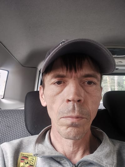 This is a medium shot, eye-level selfie of a man in his late 40s or 50s, wearing a gray baseball cap and a gray zip-up jacket with a Porsche logo on the left chest. He has fair skin, dark hair peeking out from under his cap, and some stubble on his chin and upper lip. His eyes are looking directly at the camera. The man is seated in the back of a vehicle, as indicated by the headrest of the seat in front of him and the interior of the car visible in the background. The headrest is black, and the seat beside it has a patterned fabric. Sunlight streams in from a window on the left, illuminating the interior. The background outside the window suggests a temperate climate with trees visible. The overall mood appears neutral or slightly somber. The Porsche logo on his jacket has text, including "PORSCHE" and "TRADITION". The image was taken in Zuyzino, Russia.