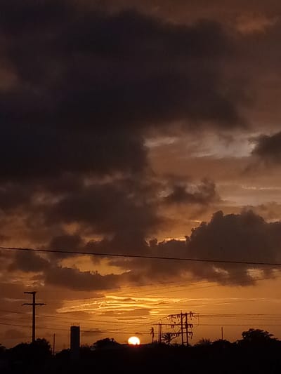 The image depicts a dramatic sunset over El Dátil, Venezuela. The sky is filled with a gradient of deep oranges and yellows, transitioning to dark, brooding grays and purples in the upper portion, indicative of heavy cloud cover. The sun, a bright semi-circle, is visible near the horizon on the right side of the frame, casting a warm glow through breaks in the clouds. Silhouetted against this vibrant sky are the faint outlines of trees and structures, including utility poles with visible wires, a tall, slender tower-like structure, and a more complex electrical transmission tower with multiple crossbars. The foreground is a dark, indistinct silhouette of the landscape. There are no people or animals visible in the image, and no actions or interactions are taking place.