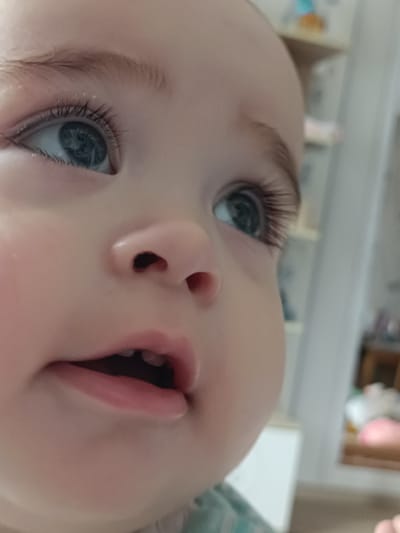 The image is a close-up, eye-level shot of a baby's face, focusing on the left eye, nose, and mouth. The baby has large, light blue eyes with long eyelashes and light brown eyebrows. The skin is fair and smooth. The baby's mouth is slightly open, revealing two small, emerging teeth. The background is blurred, showing hints of a domestic environment with shelves and what appear to be toys or decorative items. The image is likely taken indoors. The provided location of Усть-Тунтор, Russia, is a detail that cannot be determined from the visual information in the image itself.