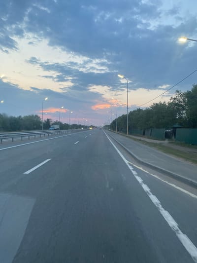 The image depicts a highway with streetlights and a cloudy sky at dusk. The road is asphalt with white lane markings. There are trees and some structures visible along the right side of the road. A blue truck is parked on the left side of the road. The sky displays shades of blue, gray, and pink, indicating the sun is near the horizon. The photograph appears to have been taken from a moving vehicle. The location is identified as the highway in Kenjekol, Kazakhstan.