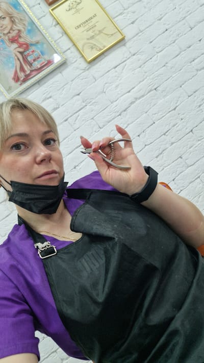 A woman with blonde hair and a black face mask stands in front of a white brick wall. She wears a purple shirt and a black apron. She holds a pair of nail clippers in her left hand, which has a black wristband. On the wall, there's a certificate in a gold frame and a framed picture of a blonde woman in a red dress. Based on the certificate in Russian, this photo was likely taken in the city of Otchor, Russia.