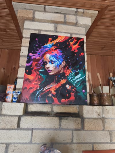 A canvas painting of a woman with vibrant, multi-colored hair and face paint is displayed on a brick mantelpiece in what appears to be a dacha or rustic indoor setting. The painting features bold, abstract splashes of color like orange, red, blue, and purple against a black background. The woman's expression is serene. To the left of the painting, a small ceramic figurine of a mouse and a box with graphics are visible. To the right, two copper pots with wooden handles are placed on the mantel. The background consists of light-colored brickwork and wooden paneling, suggesting an indoor space possibly associated with a fireplace or sauna in Востец Снт, Russia.