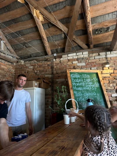 This image depicts an indoor scene, likely a small cafe or food stand, with a rustic and unfinished aesthetic. The ceiling is constructed of exposed wooden beams and planks, with some insulation visible between them. The walls are made of red brick.
In the foreground, a wooden counter runs horizontally across the frame. On the counter, there are several white paper cups and a small white basket containing wooden stirrers. A young girl with braided hair, wearing a leopard-print top, is reaching for a cup on the counter.
Behind the counter, to the left, a young man in a white t-shirt and khaki shorts is standing, looking towards the camera with a neutral expression. To his right, a white refrigerator is visible, with two cardboard boxes stacked on top of it.
On the brick wall behind the counter, a chalkboard menu is displayed. The menu is written in chalk and lists various items and prices in what appears to be Cyrillic script. Some of the items are: "Кофе - 1,5р", "Вафли 1шт - 1,5р", "Мохито - Grape - 3,5р", "Original", "Милкшейк - 5р", and "Чай - сделай себе сам".
To the right of the chalkboard, a black lamp with a visible bulb hangs from the ceiling, illuminating the menu. Green plants are visible in pots near the chalkboard and to the right of the frame. The overall impression is of a casual, possibly outdoor or semi-outdoor establishment, with a focus on simple food and drinks. The reference to "Завалочыцкі сельскі Савет, Belarus" suggests a rural or semi-rural location within Belarus.