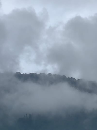 The image shows a misty mountain landscape with clouds obscuring much of the view. The top of the image is dominated by cloudy skies, with varying shades of gray and white suggesting dense cloud cover. Below the sky, a ridgeline of a mountain is faintly visible, appearing dark and silhouetted against the lighter clouds. Patches of mist or fog swirl around and over this ridgeline, partially obscuring the mountain's features. Further down, the lower slopes of the mountain are even more enshrouded in thick, diffuse fog, with only faint outlines of trees or vegetation visible as dark shapes within the haze. The overall impression is one of a damp, overcast day in a mountainous region, possibly at a higher elevation where fog is common. The scene is devoid of any human presence or specific landmarks, offering a generalized view of a foggy mountain environment.