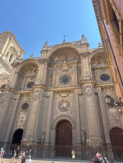 The image captures a low-angle, full-shot perspective of the grand facade of the Granada Cathedral in Granada, Spain. The sun shines brightly, casting clear shadows and highlighting the intricate stone carvings and architectural details of the Renaissance-style building. The facade features multiple arched entrances, ornate sculptures, and circular windows. A tall bell tower is visible to the left, and a glimpse of a neighboring building with a pinkish facade is seen on the right. The sky is a clear, deep blue.
In the foreground, a group of tourists are gathered on steps leading up to the cathedral. They are casually dressed in summer attire. Some are walking, others are standing and looking at the architecture. The interactions between the people appear to be casual, typical of tourists exploring a landmark. The environment is an urban setting, with the cathedral dominating the scene. The overall atmosphere is one of a bright, sunny day in a historically significant European city.