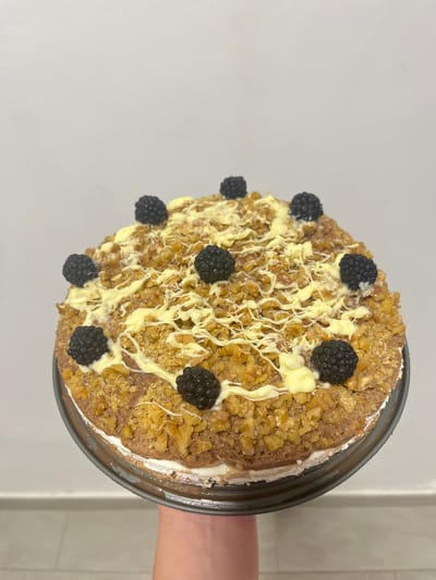 The image displays a round cake, viewed from a slightly overhead angle, held by a hand against a plain light background. The cake's top is generously covered with finely crushed, light brown nuts, providing a textured surface. Intricate drizzles of white or very light yellow frosting are patterned over the nuts. Eight dark blackberries are symmetrically arranged on the cake, offering a contrasting color and visual appeal. The cake sits in a dark, metallic round pan or serving dish, with some white frosting or cream visible around its edges. A light-skinned hand is visible on the right side of the frame, holding the cake pan. The background is a smooth, light grey surface, indicating an indoor setting with bright, even lighting. No people's faces or visible text are present in the image. The scene suggests the presentation of a carefully prepared dessert.