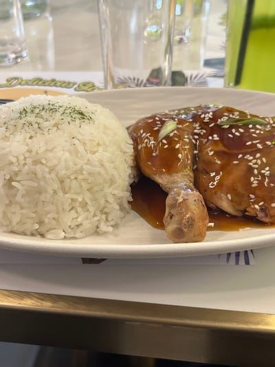 The image shows a close-up view of a plate of food, likely taken in a restaurant. The dish consists of a serving of white rice, lightly sprinkled with green herbs, and a piece of glazed chicken, possibly a quarter leg, covered in a dark sauce and sesame seeds, with some chopped green onions as garnish. The food is presented on a white plate, which rests on a table. In the background, out of focus, are glassware, possibly water glasses, and a green beverage. A portion of a placemat with a pattern is visible beneath the plate. The overall impression is of a meal ready to be eaten, with a casual dining ambiance suggested by the background elements. The specific location in Koronadal City, Philippines cannot be determined from the image alone.