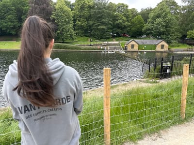 The image depicts a rear view of a person wearing a grey hooded sweatshirt with text on the back, "AVANT GARDE DES ESPRITS CREATIFS DESIGNED IN FRANCE Paris." The person is standing on a path next to a body of water, likely a lake or river, with a grassy embankment and a fence in the foreground. In the background, there are trees, a building with a green door, and several swans on the water. The scene appears to be a park or recreational area. Without further context or specific landmarks, pinpointing the exact location within Leeds is not possible, however, the presence of a lake, swans, and parkland suggests a location like Roundhay Park or Temple Newsam Park.