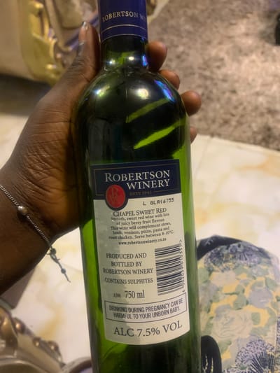 In Akpajo, Nigeria, a dark-skinned individual's hand, adorned with a silver-colored chain bracelet, holds a green glass bottle of Robertson Winery Chapel Sweet Red wine. The bottle's label, marked "ESTD 1941," details the wine as "Smooth, sweet red wine with lots of juicy berry fruit flavour," to be served between 8-10°C, and notes it "CONTAINS SULPHITES." The volume is specified as "750 ml" with an "ALC 7.5% VOL," and a warning states, "DRINKING DURING PREGNANCY CAN BE HARMFUL TO YOUR UNBORN BABY." A batch number, L GLA16755, and a partial barcode are visible. The blurred background suggests an indoor setting, with elements including a light-colored textured surface, a floral-patterned fabric, and a decorative object of a light, possibly golden, hue.