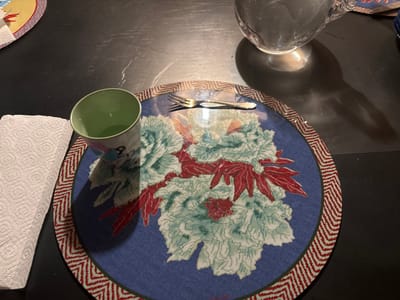 The image shows a table setting in a private residence in Celerina/Schlarigna, Switzerland (46.514, 9.859). It features a round placemat with a blue background and red and white chevron border. The placemat displays a floral pattern in red and green hues. A fork lies on the placemat. A green glass, filled with a clear liquid, is placed on the placemat. A white paper napkin lies on the table next to the placemat. A clear glass pitcher with a handle, filled with water, sits on the table, above and to the right of the placemat. The table surface is dark grey. The scene is empty of people. The lighting is soft and warm.