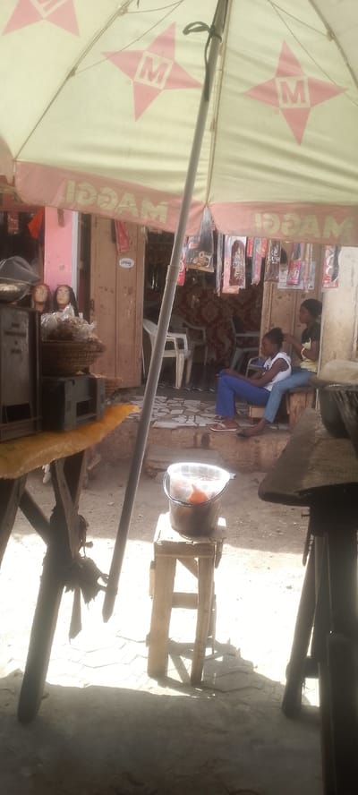 The image portrays an outdoor commercial scene in Katsina, Nigeria, near Muhammadu Dikko Stadium. A large, light-colored umbrella with red "M" logos and "MAGGI" branding (visible in reverse) provides shade in the foreground, supported by a central pole.
Two young females are seated in the mid-ground. The female on the left wears a white top and blue pants, sitting on a low surface. The female on the right, in a dark top and light blue jeans, sits on a wooden bench, actively braiding or styling the hair of the first individual.
To the left foreground, a wooden table supports dark, rectangular objects and a woven basket containing items in clear plastic bags. Behind this table, two mannequin heads with dark hair are visible. Directly beneath the umbrella, a clear plastic bucket containing a reddish-brown substance rests on a small wooden stool.
The background features the entrance to a shop, characterized by a partially open wooden door with a white label reading "LIFTING." Inside the shop, white plastic chairs are visible, and numerous packaged hair extensions hang from an overhead support. A patterned reddish fabric is also visible within the shop's interior.
The ground surface in the foreground is unpaved, transitioning to a patterned tiled area near the shop entrance. Strong sunlight creates distinct shadows, indicating an outdoor, daytime setting. The adjacent shop wall to the left appears to be painted a pinkish hue.
