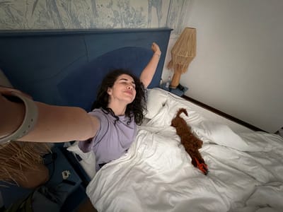A young woman with curly dark hair is stretching in bed, taking a selfie. She is wearing a light purple t-shirt and has a bracelet on her left wrist. Her eyes are closed, and she appears to be waking up. The bed is covered with white sheets and a duvet. To the right of the woman, a small brown dog is lying on the bed, with its head resting on a toy. The bedroom has a blue headboard with a textured wallpaper featuring a nature-inspired pattern behind it. A bedside table with a lamp and a bottle of water is visible to the right. This scene is located in a bedroom in the city of Banya, Bulgaria.