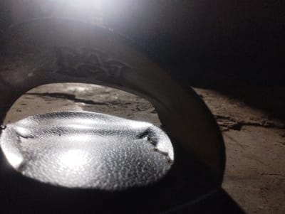 This image, taken in Kubare, Nigeria, depicts a dimly lit, confined environment. In the immediate foreground, a dark, semi-reflective object with a distinct granular or pebbled texture occupies the lower frame, exhibiting several minor indentations and bright reflections from an overhead light source. An arch-shaped aperture is visible above this object, framing a view of cracked, uneven ground or flooring in the background, which appears dry and dusty with variations in tone. The surrounding structure forming the arch is dark and curved, suggesting an enclosed space such as the interior of a large pipe, a drum, or a vehicle component. No individuals or distinct actions are present.