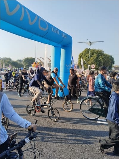 This detailed image captures an outdoor public event on an asphalt road in Sragen, Indonesia, under a clear, bright blue sky with evident shadows suggesting a sunny morning or late afternoon.
In the upper left and central portions of the frame, a large, inflatable blue archway spans the road. The arch is branded with "VIVO" in white/light gray text on its left side and "ELITE CELL" in similar styling on its right side. Smaller, partially visible text below "VIVO" reads "Smart...", while "Smart..." is also visible vertically on the right pillar of the arch.
The scene is densely populated with numerous individuals, primarily adults, many of whom are riding bicycles. In the foreground, from the left, a person's light blue long-sleeved arm and part of their torso are visible, holding the handlebars of a bicycle. In the central foreground, a woman wearing a dark blue hijab, a dark blue long-sleeved shirt, and light-colored pants is actively riding a dark-colored folding bicycle, smiling slightly towards the right. To her immediate right, a man in a light-colored baseball cap, a light blue vest over a dark shirt, and khaki shorts is also riding a bicycle. Further right, a man in a black cap and a shiny blue long-sleeved shirt is pedaling a large dark green bicycle with a prominent rear fender. Another person in a blue hoodie and dark pants is partially visible walking on the far right foreground.
Various types of bicycles are present, including several folding bikes with small wheels and mountain bikes. In the mid-ground, a man in a black t-shirt and dark pants, wearing light blue shoes, is walking alongside his black bicycle. Behind this man, a vendor holds a large cluster of colorful balloons.
The background features several large, green leafy trees, indicating a well-vegetated urban area. To the right of the archway, distinctive horn-shaped sculptures, likely part of a fountain system, are visible, with water spray faintly discernible. These sculptures are characteristic of the Tugu Gading or Taman Krido Anggo area in Sragen. A decorative streetlight with ornate metalwork is positioned above the trees on the upper right. The asphalt road surface shows minor wear, and a white lane line is visible on the right side. Long shadows of people and bicycles extend across the road, indicating the direction of sunlight. The overall atmosphere suggests an active community gathering or organized cycling event.