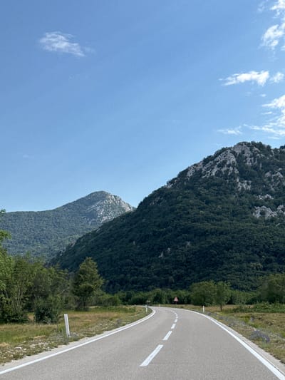The image captures a scenic view of a winding road leading through a mountainous landscape. The road, paved and marked with white lines, curves gently to the left, disappearing into the distance. On either side of the road, the terrain is covered with green vegetation, including trees and grasses. In the background, majestic mountains rise, their slopes densely forested. The sky above is a clear blue, with a few wispy white clouds scattered across it. The overall impression is one of natural beauty and tranquility. The location is specified as the city of Dragalj, Montenegro. There are no people or animals visible in the image. Essential details of the scene include the road, mountains, vegetation, and sky.