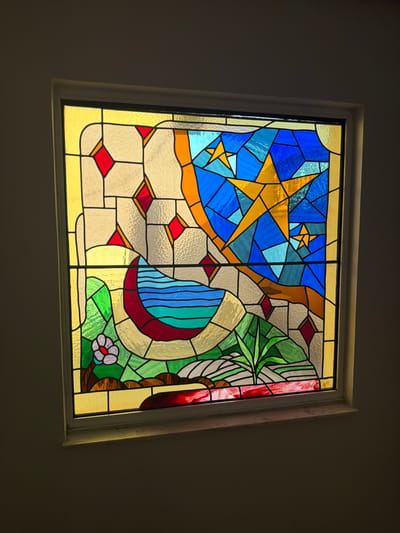 The image shows a stained glass window set into a wall. The window is rectangular and framed by a white border. The stained glass depicts a stylized landscape with celestial elements. In the upper portion, a bright yellow star is prominent against a background of various shades of blue, representing the night sky. There are other smaller stars and geometric shapes in blue and yellow.
The lower part of the window features a crescent moon shape in deep red, filled with wavy lines of blue and green, suggesting water. To the left of the moon, there is a single, stylized flower with white petals and a red center, growing from a brown stem and green leaves. The ground around the flower and the moon is depicted with patches of green and earth tones, interspersed with white and grey abstract shapes. There are also red diamond-shaped pieces of glass scattered throughout the scene. A thin wooden windowsill is visible below the stained glass window. The surrounding wall is painted a dark, neutral color, creating a contrast with the illuminated window. The lighting suggests the image was taken indoors, possibly during the day as light passes through the stained glass. The location in Arcahueja, Spain, is not evident from the visual information provided in the image.