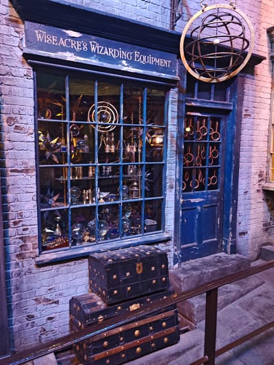 The image depicts the exterior of "Wiseacre's Wizarding Equipment" shop, located in Watford, United Kingdom. The shop facade is made of aged brick, painted in shades of blue and grey. A large display window with a grid of blue-painted bars showcases an array of magical items, including orbs, telescopes, and other scientific instruments. Above the window, a sign reads "Wiseacre's Wizarding Equipment" in stylized lettering. To the right of the window, a blue door features a smaller window with ring-shaped decorations. Hanging above the door is an ornate, spherical metal sculpture. Two black trunks, decorated with brass studs, are stacked on the step in front of the shop. A metal railing runs along the edge of the sidewalk in front of the store. The overall scene suggests a whimsical and theatrical setting.