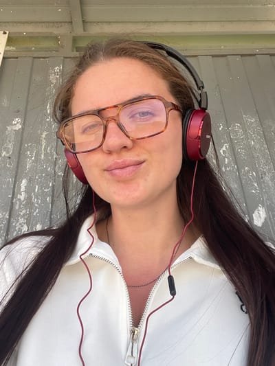 A woman is seen in a selfie, turned to the left in the frame. She wears headphones and glasses, and a white zipper jacket. The background consists of a grey, weathered surface, possibly a wall or floor. No specific details point to Molokovo, Russia.