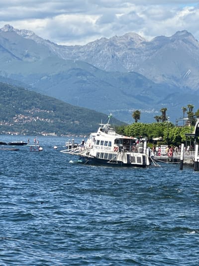 The image depicts a ferry docked at a pier on a body of water, with a mountainous landscape in the background. The ferry, named "FRECCIA DEI GIARDINI," is white with multiple windows and appears to be carrying passengers. Several people are visible on the pier, some boarding or disembarking the ferry. The water is choppy, with small waves. In the distance, a hillside town with red-tiled roofs is visible against a backdrop of lush green trees and imposing mountains under a cloudy sky. The scene is set in San Giovanni, Italy, likely on a lake given the mountainous terrain and ferry operation.