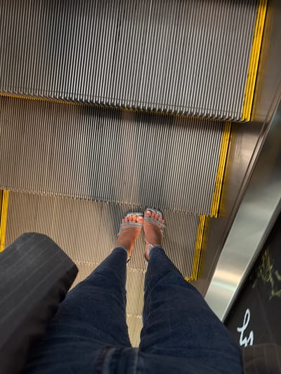 A person stands on an escalator in Vnukovo, Russia. The escalator steps are made of metal with yellow safety strips along the edges. The person is wearing blue jeans and light blue jeweled sandals. A dark-colored bag is held in the person's left hand. The background shows the metal and yellow components of the escalator.