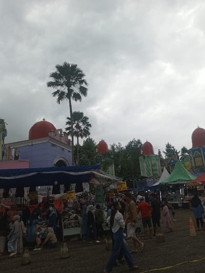 This image depicts an outdoor market or festival occurring in Suddepalle, India, under an overcast sky. The environment is characterized by a dark paved ground surface featuring concrete bollards and a yellow demarcation line. Tall palm trees are visible against the cloudy sky, alongside other dense green foliage in the background.
Architecturally, the scene includes multiple structures with distinct red dome-shaped roofs. A prominent building on the left is light purple with white architectural accents and a large red dome. Further back on the right, other buildings exhibit similar red domes paired with green and yellow decorative elements, suggesting a themed entertainment venue.
The foreground and midground are densely populated with numerous individuals. People are engaged in walking, standing, and browsing among various market stalls. These stalls are covered by blue, green, and white tarpaulin canopies, displaying merchandise such as clothing and other goods. Signage on some stalls indicates prices, for example, "50.000" and "50,000". Many individuals, particularly women, wear head coverings. A young woman in a white long-sleeved shirt and blue jeans walks towards the right in the foreground, with a man in a brown shirt and shorts walking beside her. Multiple other adults and children are visible throughout the crowded market area, with some individuals appearing to examine items on display at the stalls. An orange traffic cone is present on the far right.