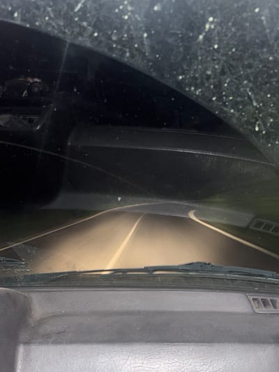The image is taken from the interior of a vehicle, looking through the windshield at a road at night. The windshield is dirty with numerous white specks, suggesting it's covered in dust or dried liquid. The headlights of the car illuminate a paved road with a white dashed line down the center. The road curves to the right, and there are faint reflections on the road surface. The dashboard of the car is visible in the foreground, with textured plastic and a vent on the right side. The sky above is dark. The location is specified as Tsar Kaloyan, Bulgaria.